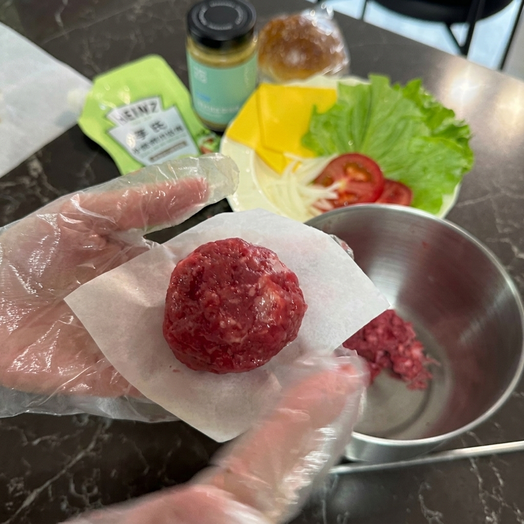 A gloved hand holding a round ball of raw ground beef resting on a small square cut of white parchment paper.