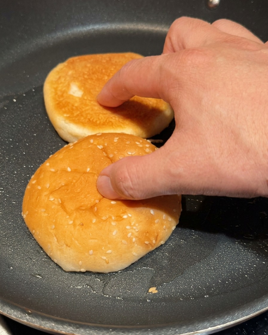 A hand lightly pressing two halves of a burger bun onto a hot black skillet to toast.