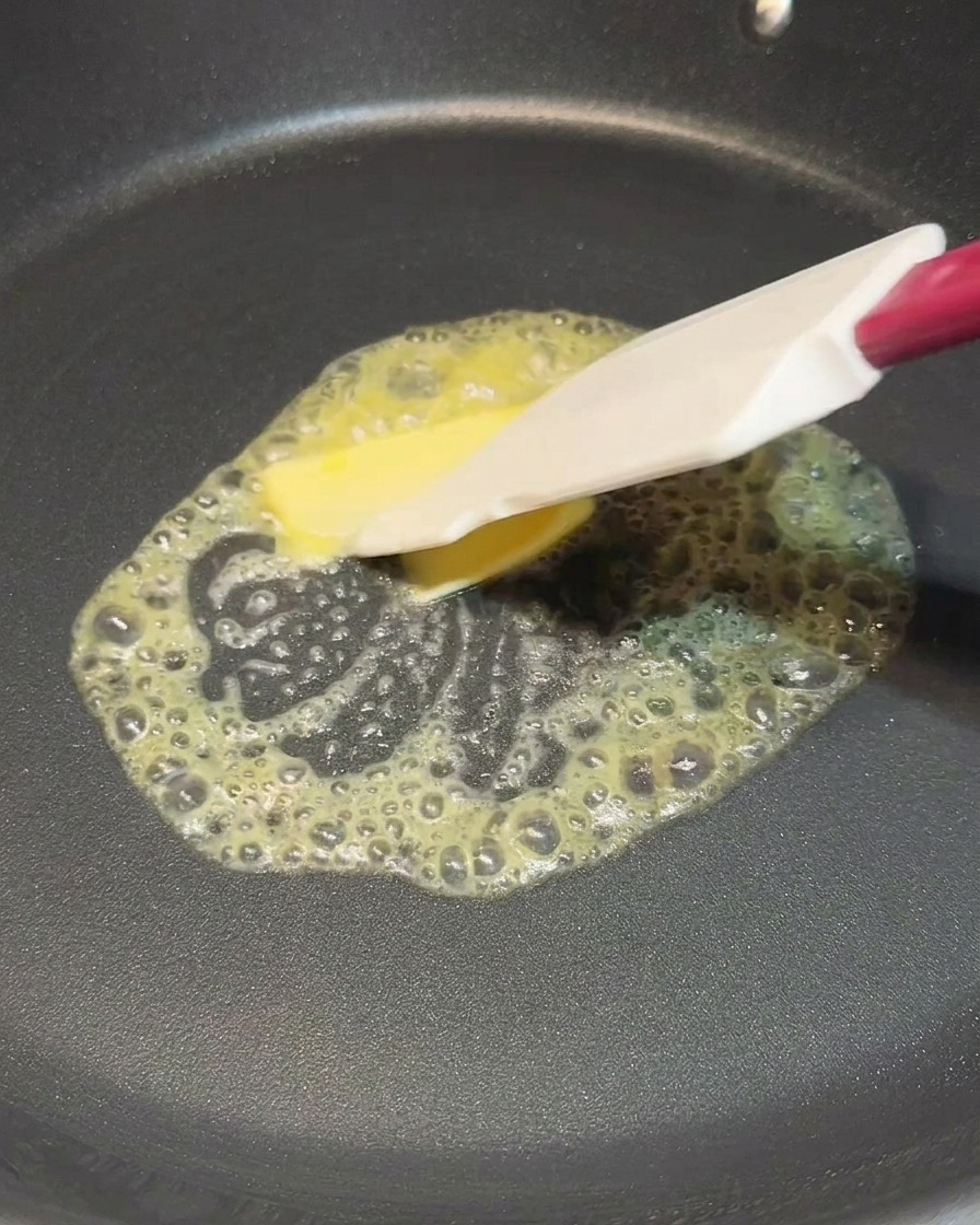 A white spatula spreading a melting square of yellow butter in a hot black frying pan.