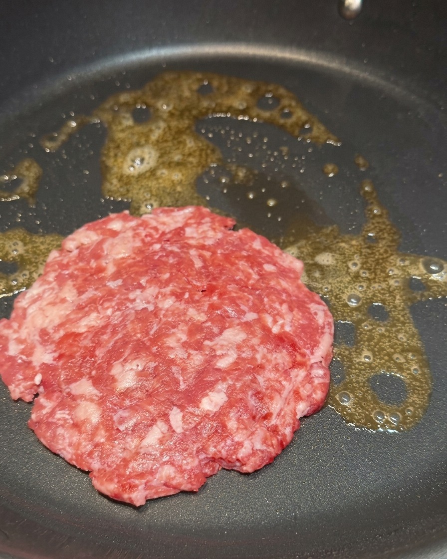 A raw, marbled beef patty sizzling in melted butter in a black frying pan.