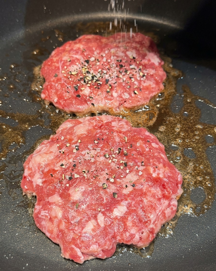 Sprinkling coarse salt and cracked black pepper onto two raw beef patties sizzling in a pan.