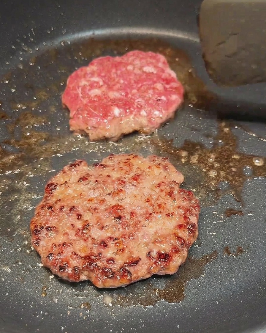 Two beef patties cooking in a pan, one recently flipped to reveal a browned crust.