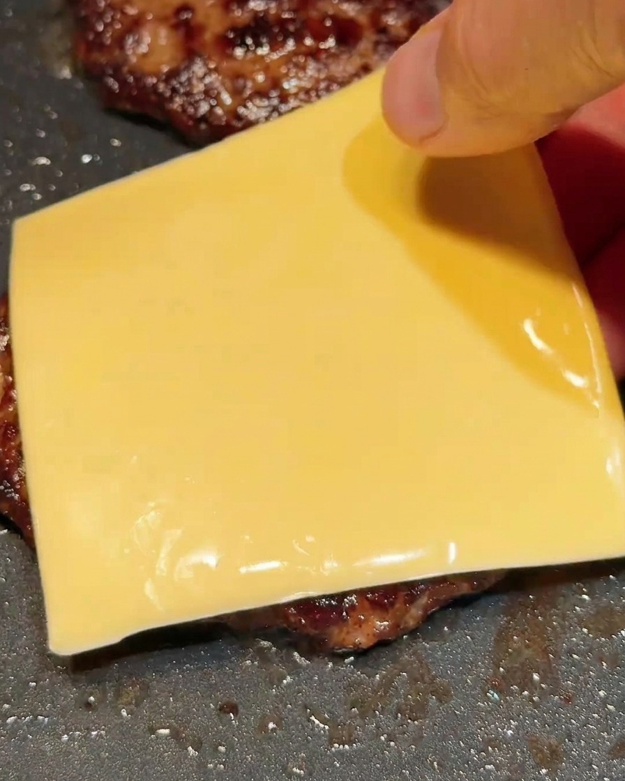 A hand placing a square slice of yellow cheese onto a hot beef patty in a skillet.