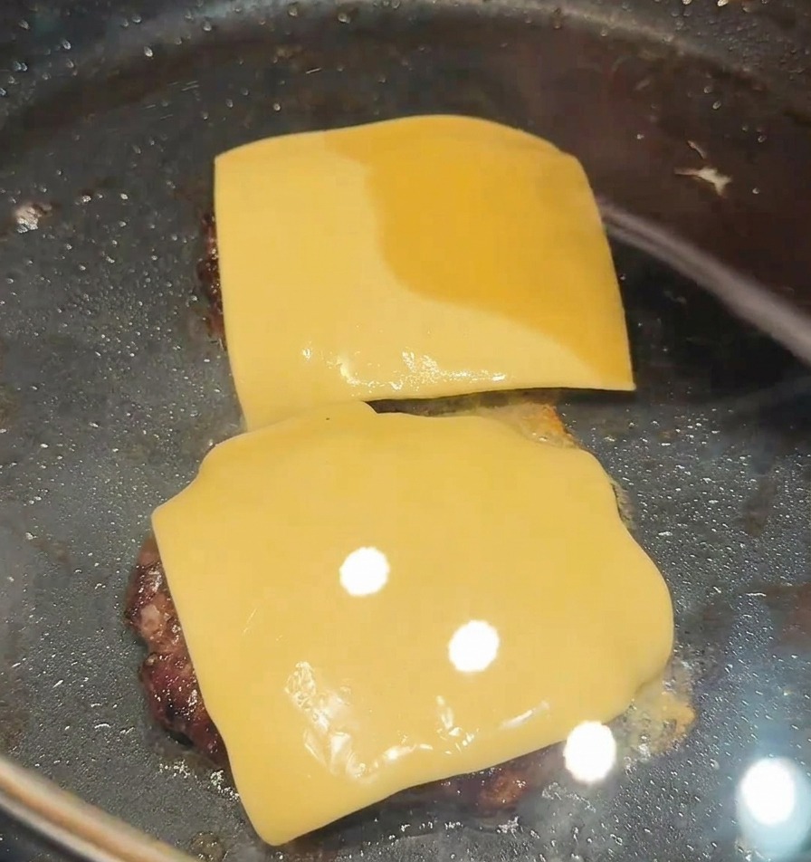 Two square slices of yellow cheese being placed on top of two searing beef patties in a black skillet.