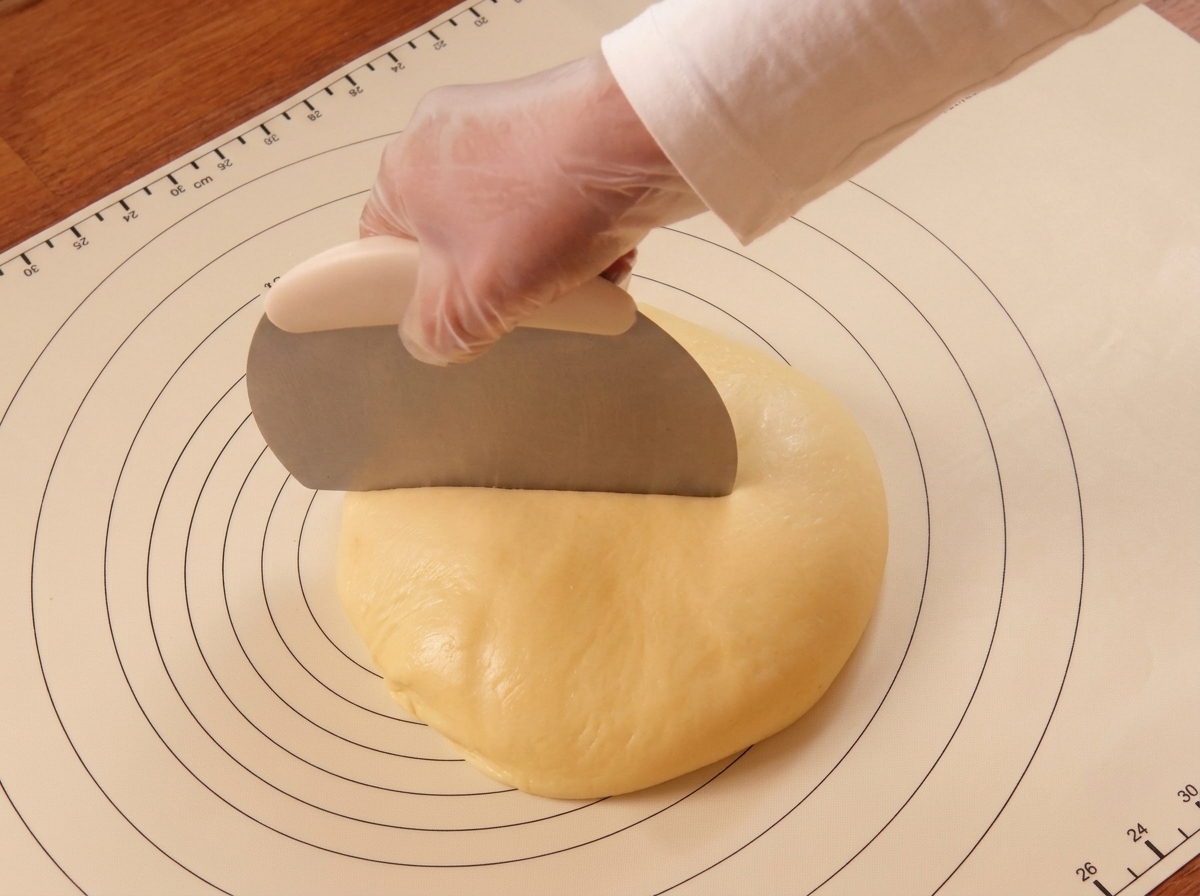 A gloved hand using a metal dough scraper to cut a mound of dough on a marked silicone mat.