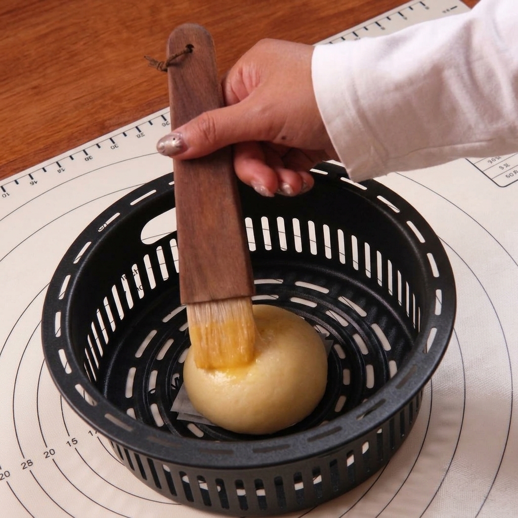 A hand using a flat pastry brush to coat a smooth, proofed dough ball with shiny egg wash.