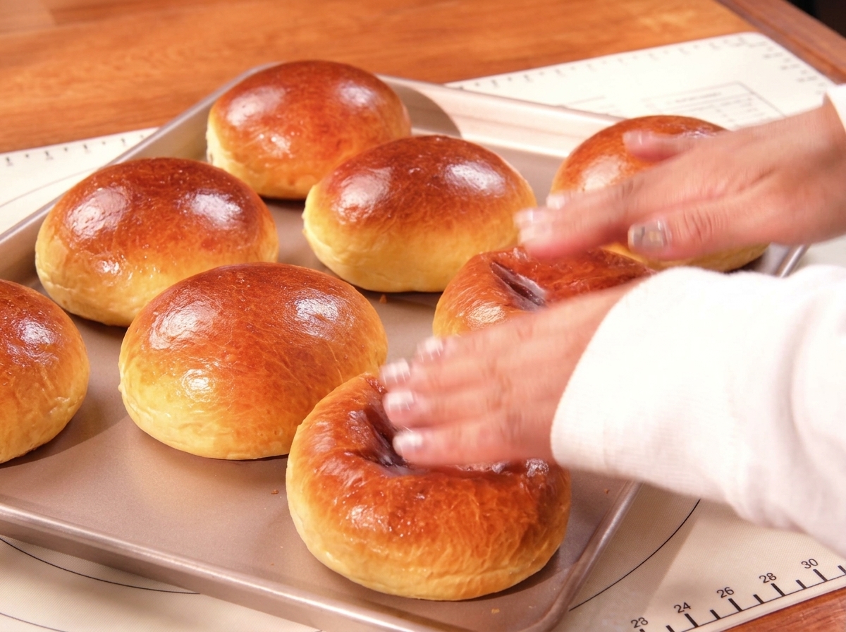 A hand gently tapping the top of freshly baked, shiny golden-brown brioche buns resting on a flat baking tray.