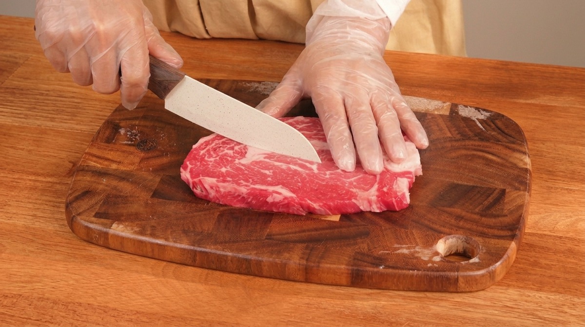 Gloved hands using a white knife to slice a thick piece of raw marbled red steak on a wooden cutting board.