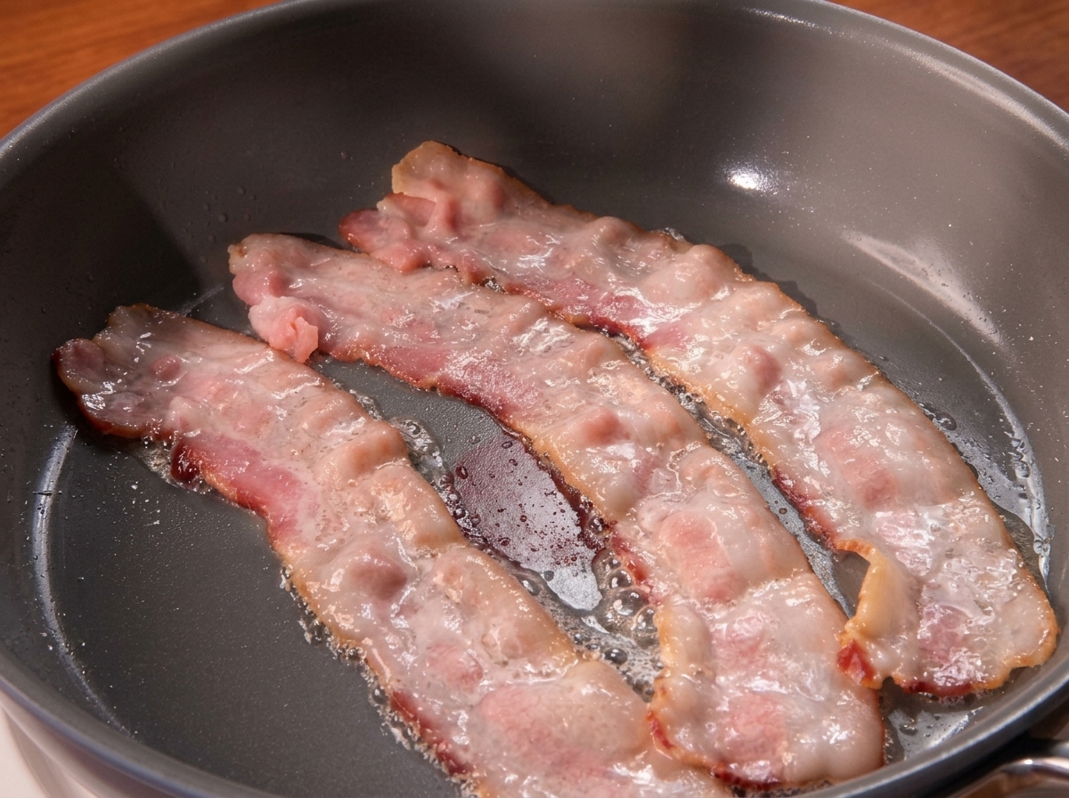 Three slices of streaky bacon sizzling and rendering their fat in a hot frying pan.
