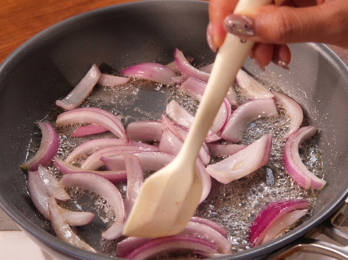 A spatula stirring softening red onion slices as they sauté in a bubbling hot pan.