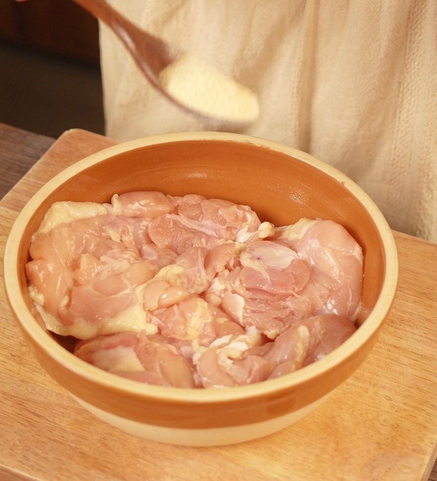 A wooden spoon sprinkling light-colored garlic powder over raw, boneless chicken thighs in a brown ceramic bowl.