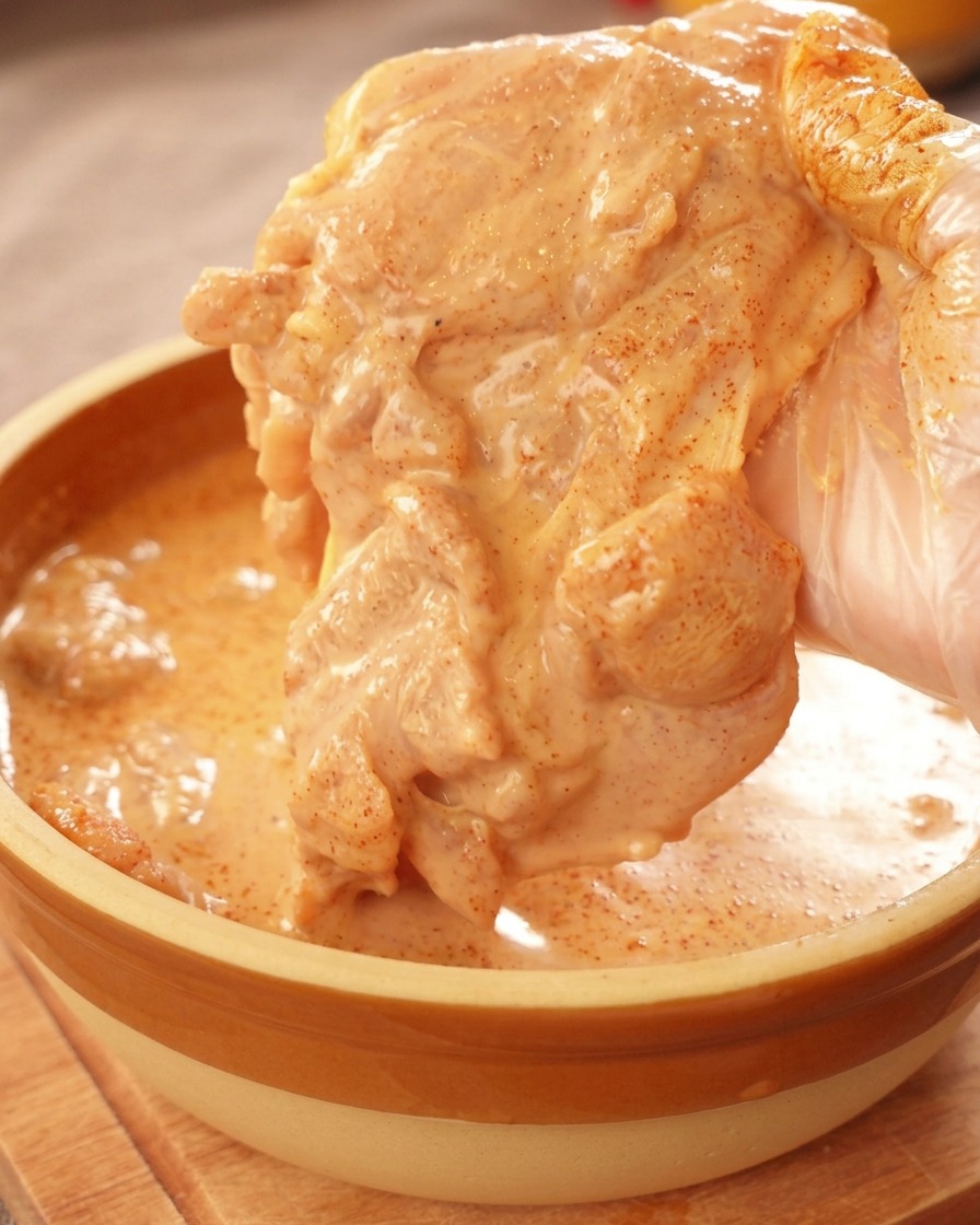 A gloved hand lifting a piece of raw chicken thigh generously coated in a thick, vibrant orange marinade.