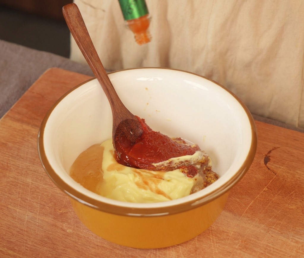 Hot sauce being squeezed into a yellow bowl containing mayonnaise, whole grain mustard, and a red paste.