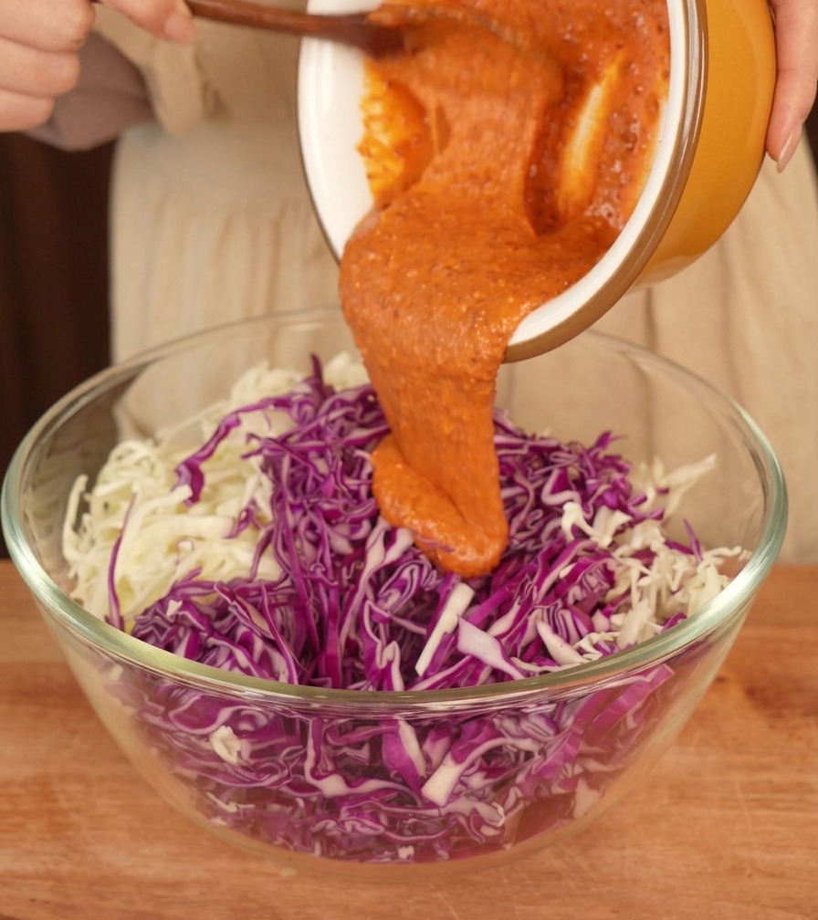 Creamy orange dressing being poured from a yellow ceramic bowl onto a mound of shredded purple and green cabbage in a glass bowl.