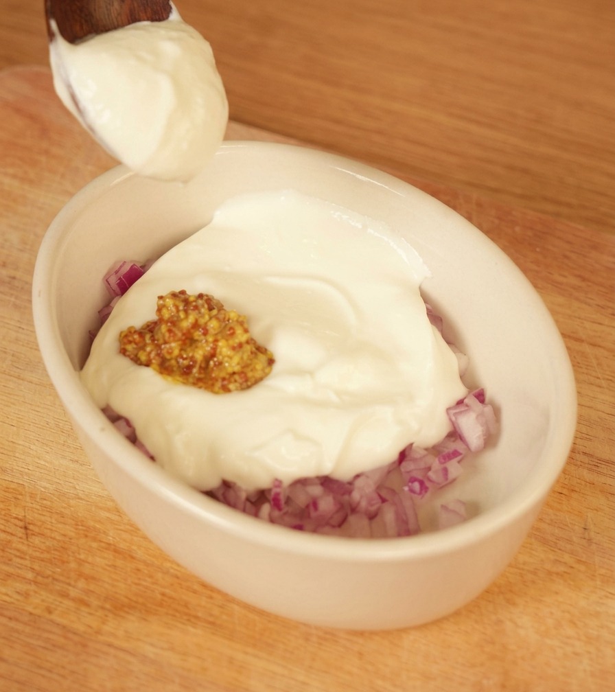 A spoonful of thick white yogurt being added to a small white bowl containing chopped red onions and whole grain mustard.