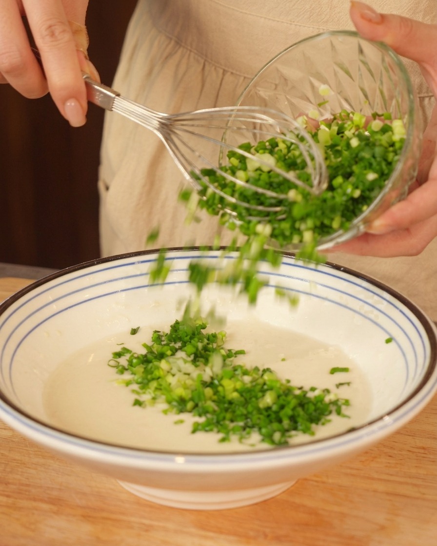 Pouring chopped green onions into a bowl of white batter while holding a whisk.