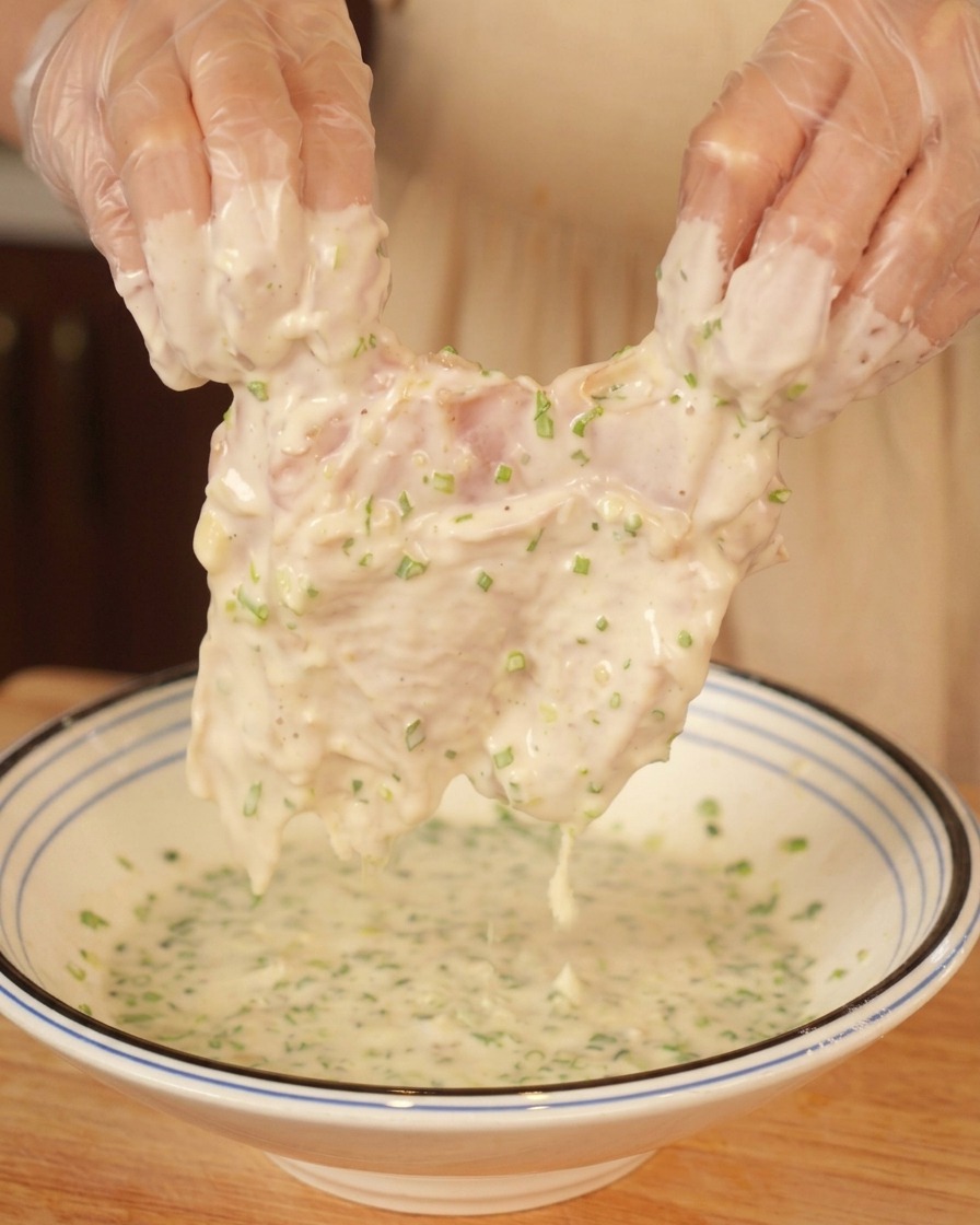Gloved hands lifting a raw chicken thigh heavily coated in a thick, green-flecked white batter.