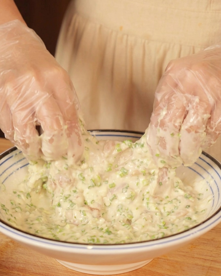 Gloved hands mixing and coating a piece of raw chicken in a bowl of thick batter.