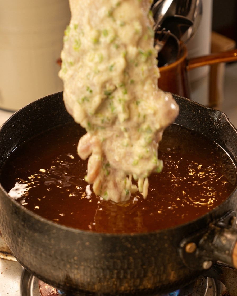 Lowering a battered chicken thigh into a pot of bubbling hot, dark oil.