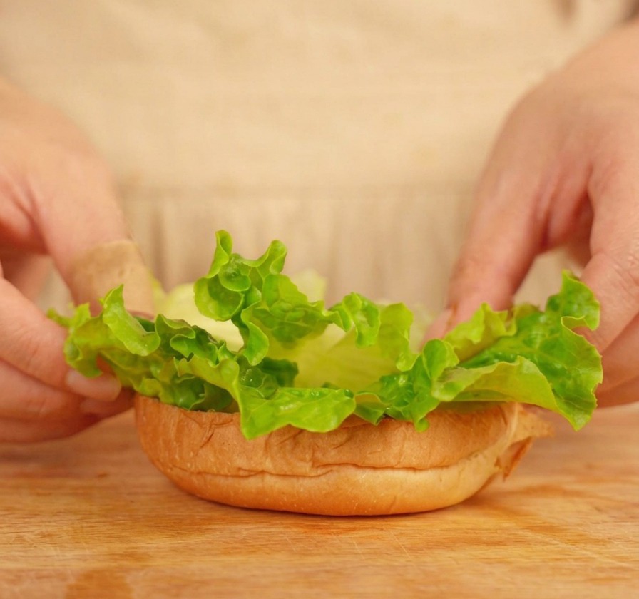 Hands carefully placing ruffled green lettuce leaves onto a toasted burger bun base.