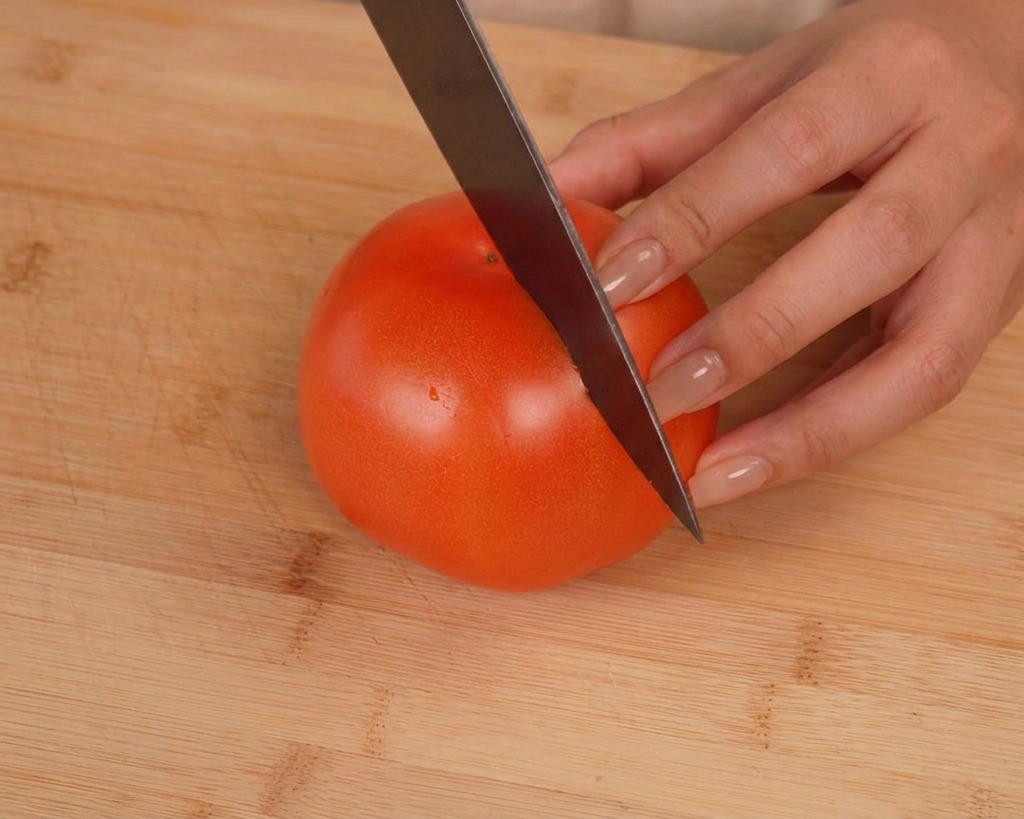 A sharp knife creating a shallow cross-shaped cut on the top of a ripe red tomato sitting on a wooden cutting board.