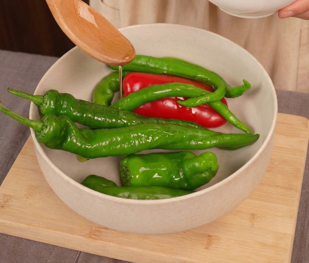 Clear cooking oil being drizzled from a wooden spoon onto a mix of fresh red and green chili peppers in a light-colored bowl.