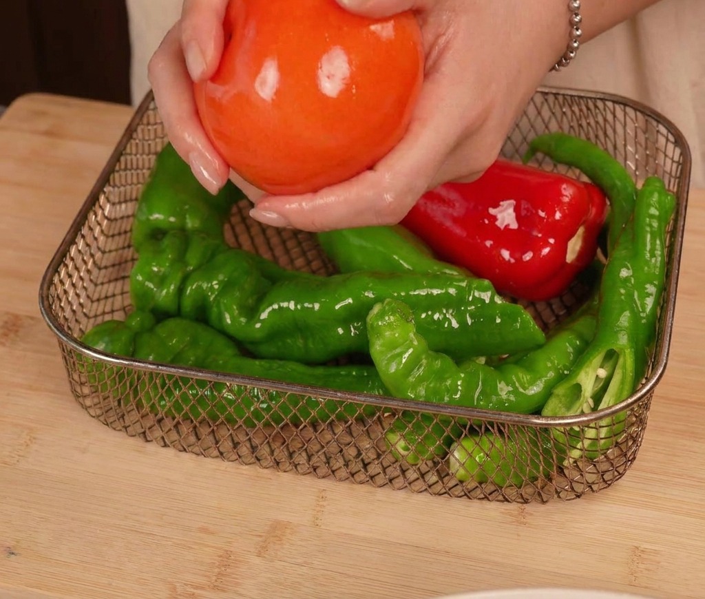 Hands placing a glossy, oiled tomato into a metal wire air fryer basket alongside several green and red peppers.