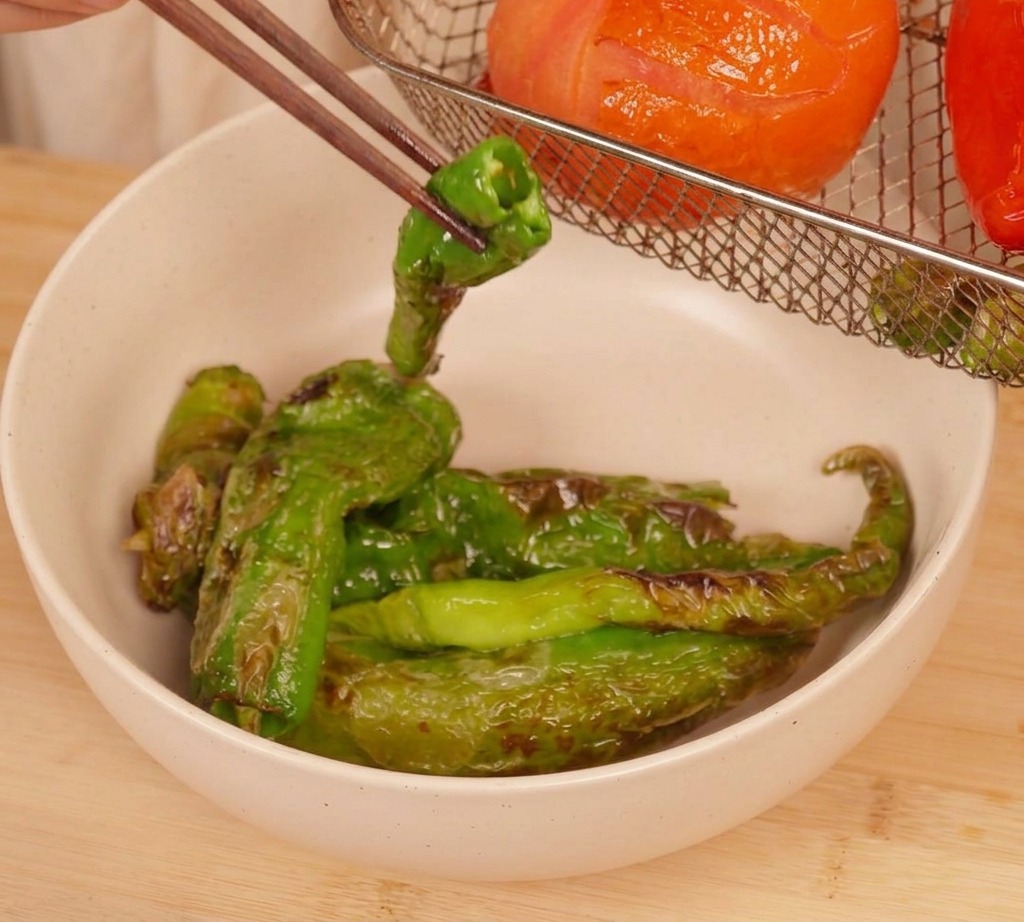 Chopsticks lifting a charred green chili pepper from an air fryer basket containing a roasted tomato and other peppers.