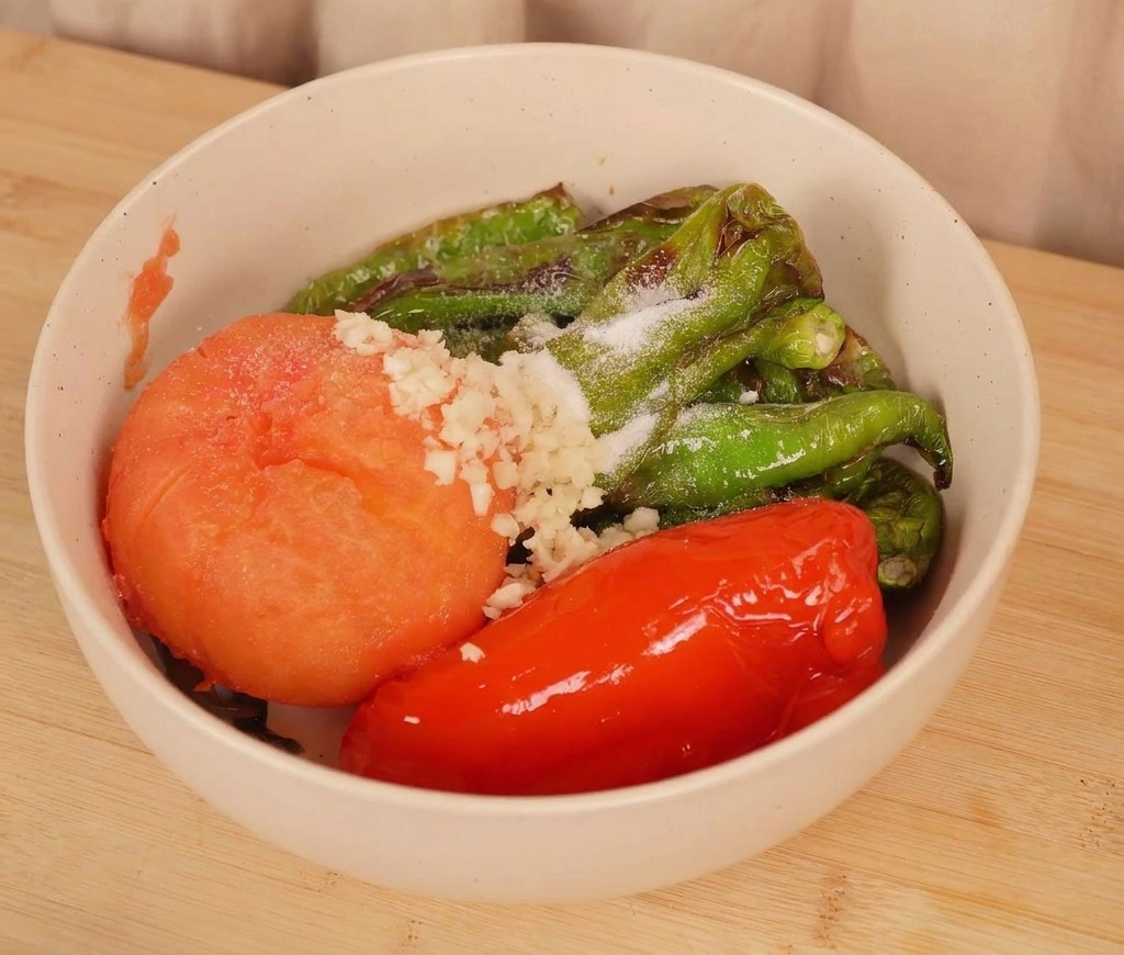 A bowl of roasted peppers and a tomato topped with fresh minced garlic and a sprinkling of salt before being mixed.