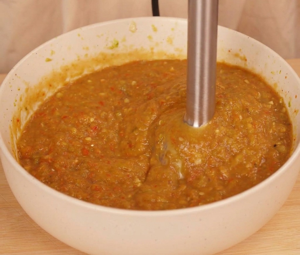 An immersion blender processing charred peppers and tomato into a thick, textured orange-brown chili sauce in a white bowl.