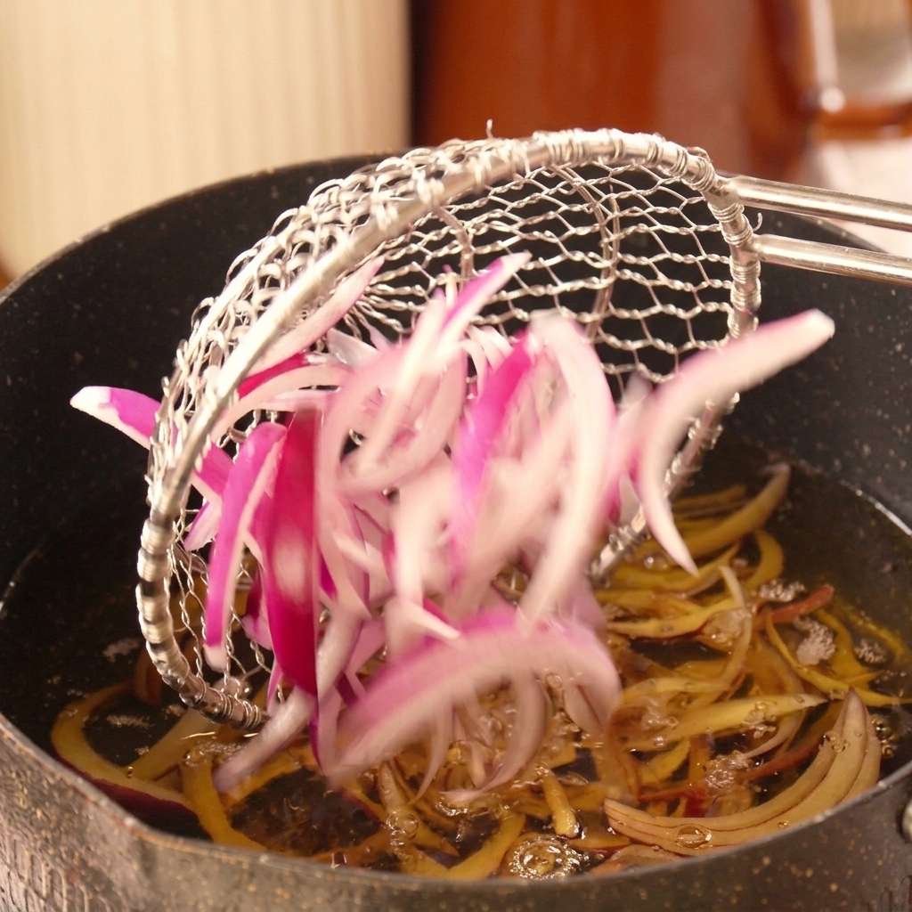 Sliced red onions being lowered from a wire skimmer into a pot of bubbling hot oil.