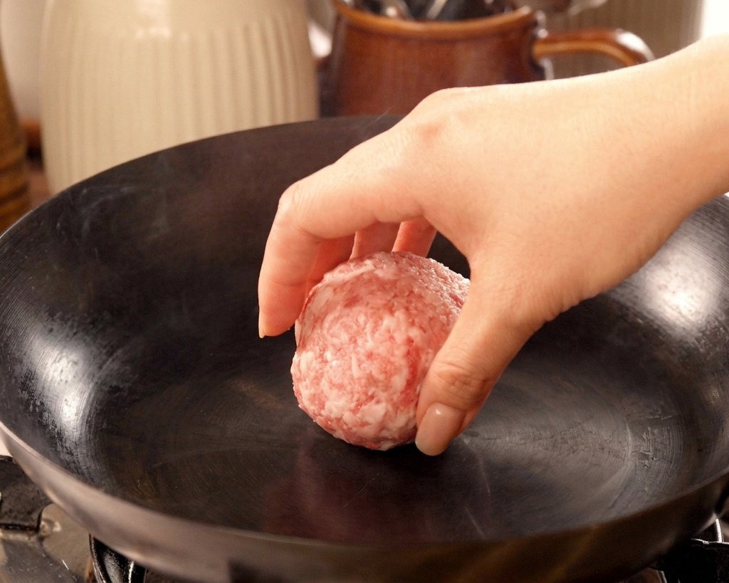 A hand gently dropping a round portion of raw ground beef into a smoking hot, ungreased cast iron wok.