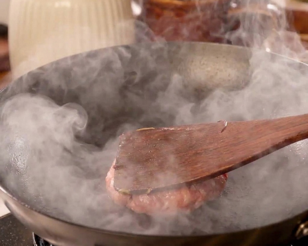 A flat wooden spatula pressing down hard on a ground beef patty, flattening it against the surface of a hot, smoking wok.