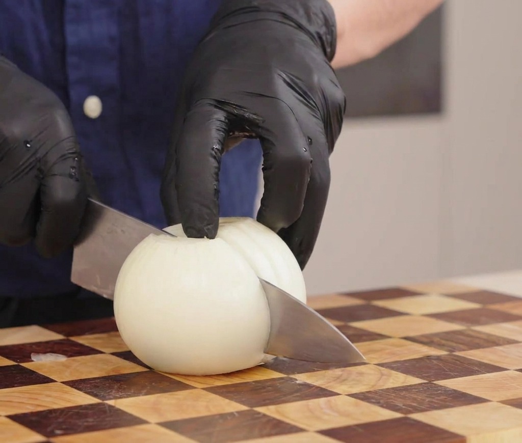 Gloved hands holding a knife to slice a white onion into strips on a checkered wooden cutting board.