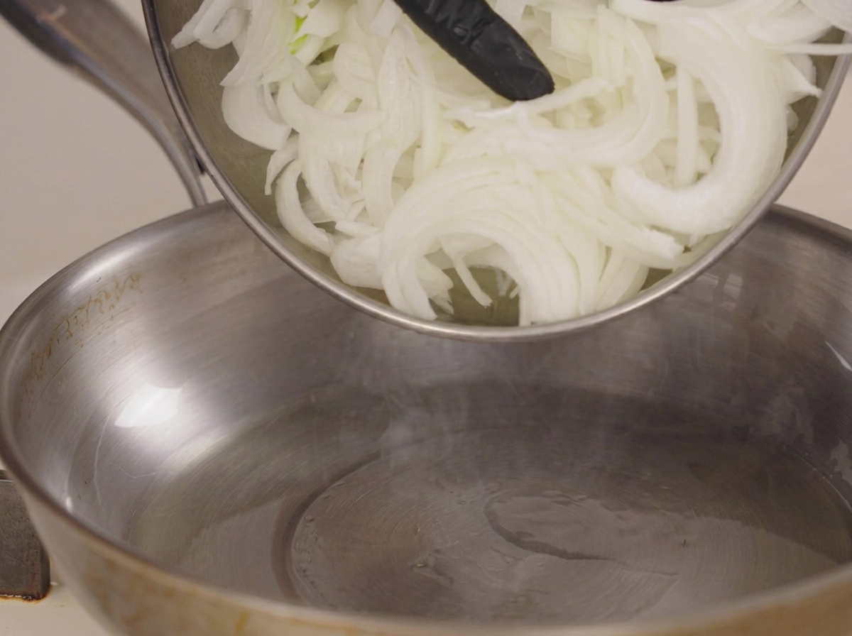 Raw sliced onions being dropped into a stainless steel pan.
