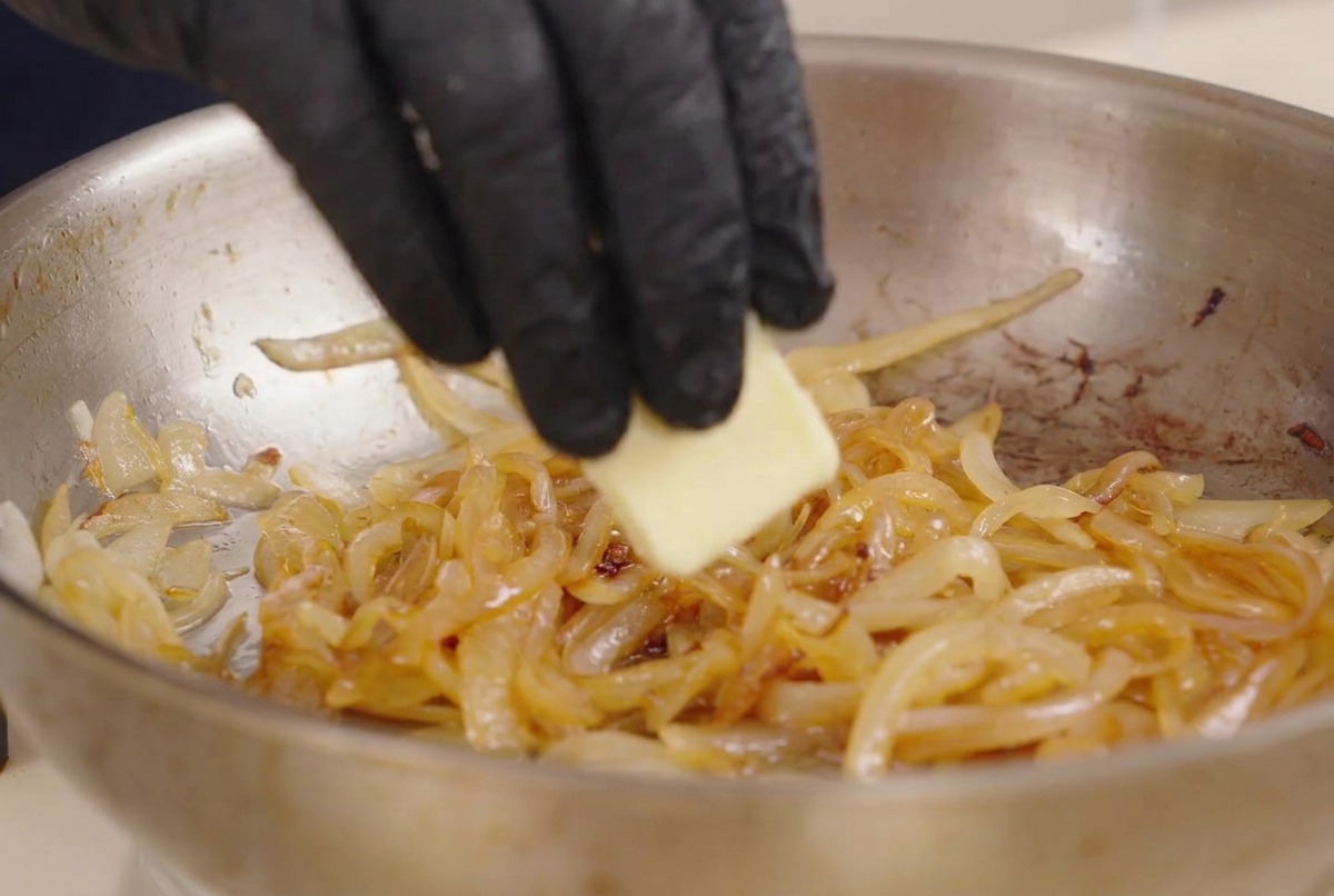 A hand adding a piece of butter into a stainless steel pan containing sautéed white onions and dark liquid seasoning.