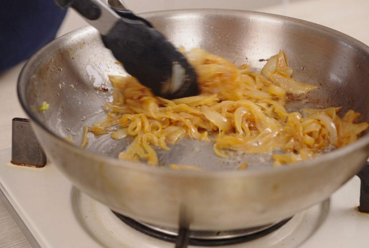 Tongs stirring deeply browned, caramelized onions in a stainless steel pan.