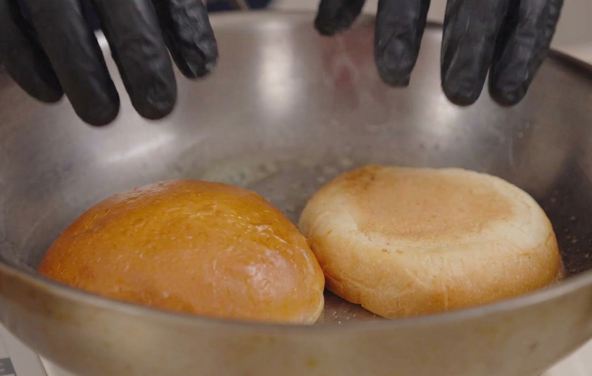 Hands in black gloves pressing two burger bun halves facedown into a hot pan with melted butter.
