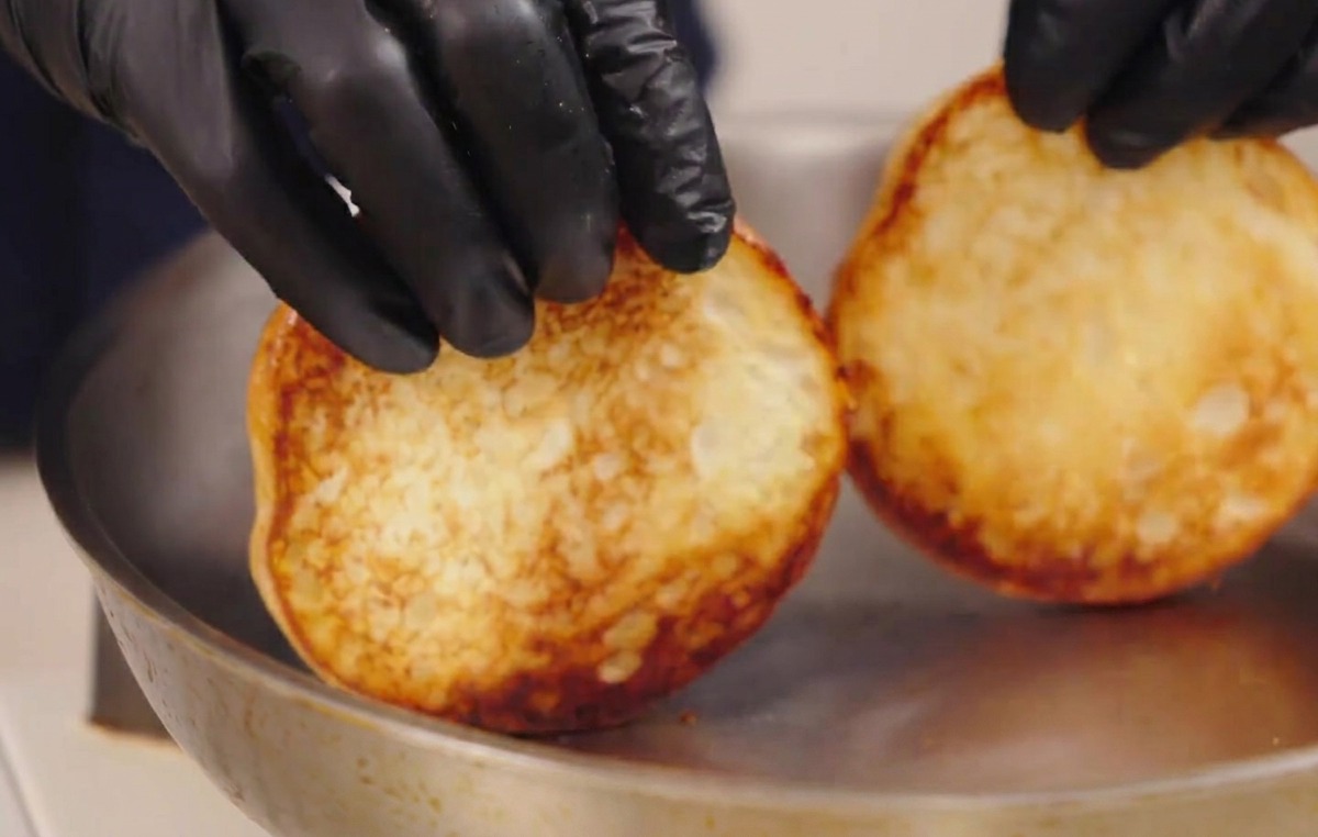 Gloved hands lifting two toasted burger buns from a pan, showing their perfectly browned, caramelized undersides.