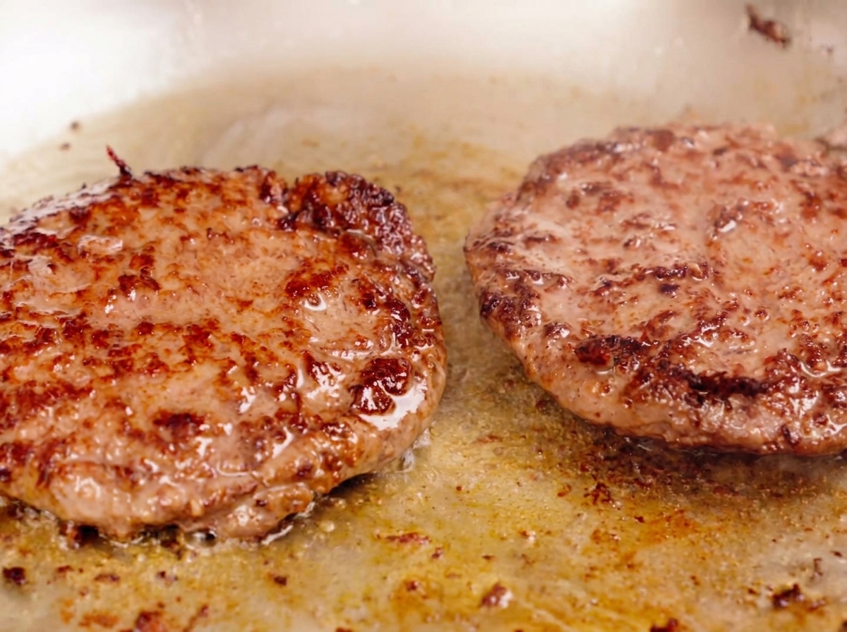 Two thick beef patties searing in a pan, with a well-developed brown crust and juices bubbling on the surface.