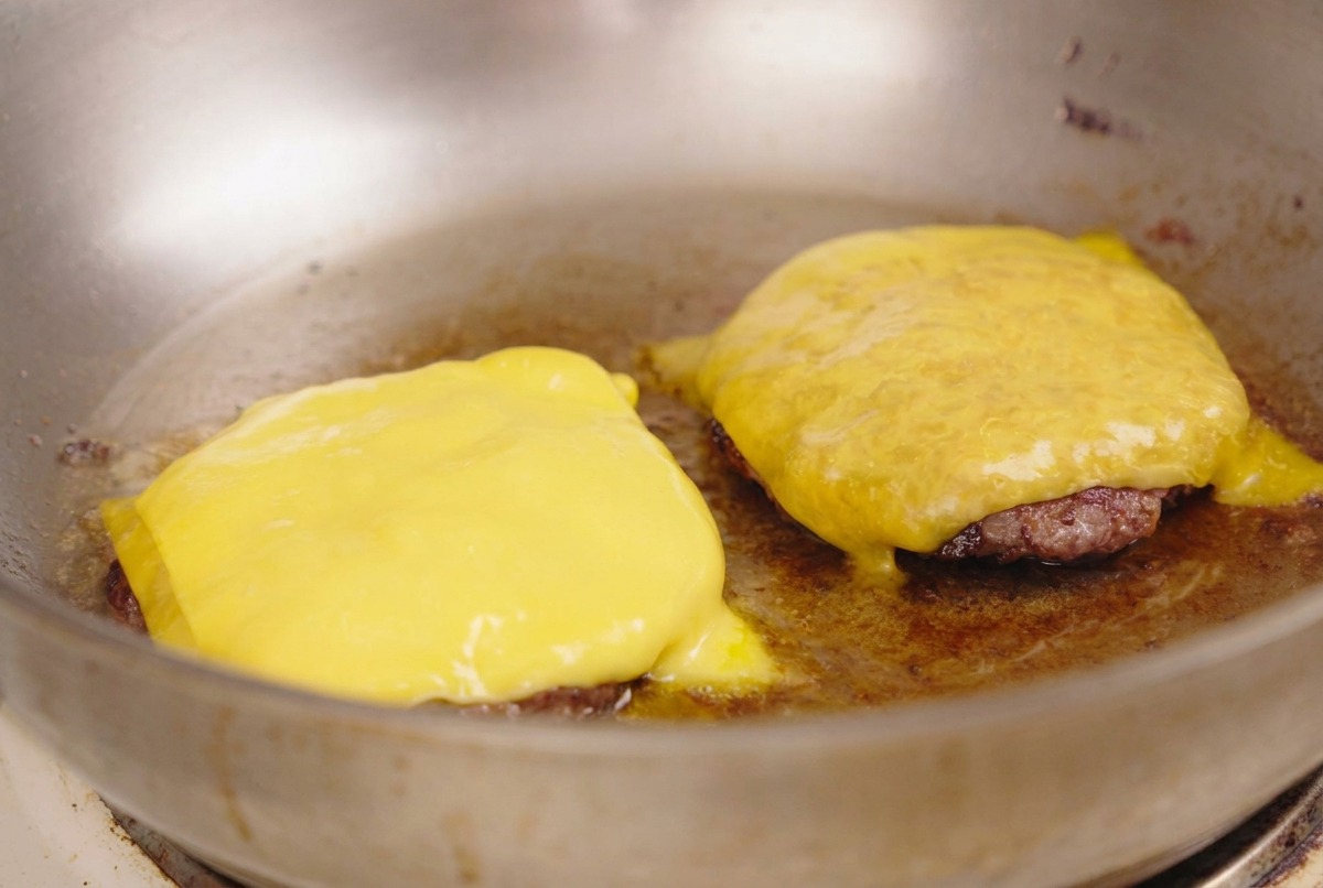 Bright yellow cheese slices melting evenly over two seared beef patties in a stainless steel pan.