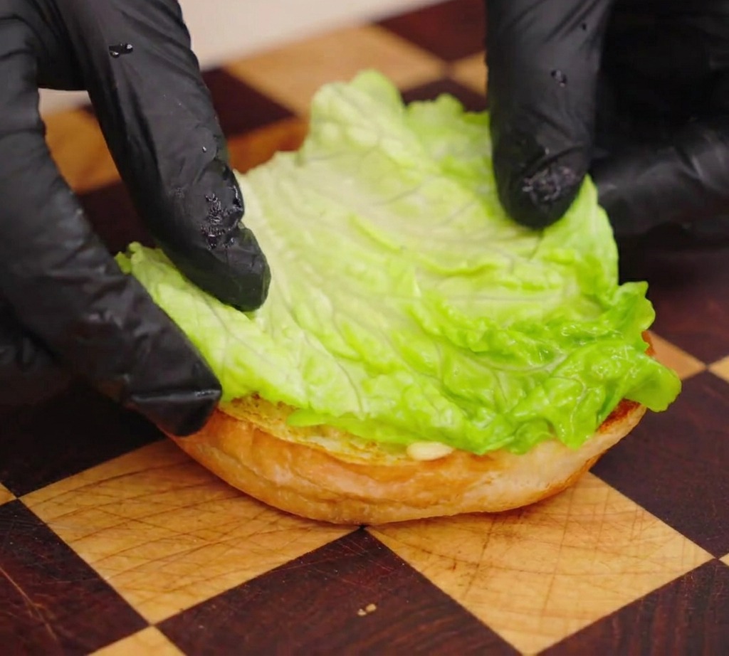A person wearing black gloves placing a bright green lettuce leaf on top of a toasted burger bun.