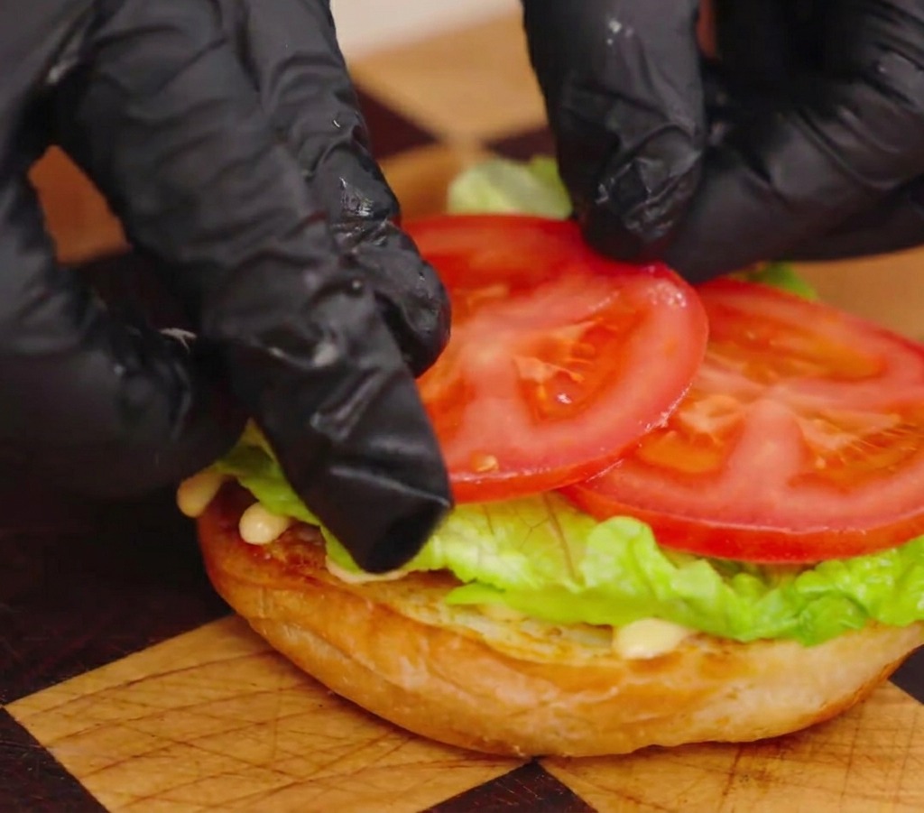 A close-up of gloved hands arranging two thick, juicy slices of red tomato onto a layer of green lettuce on a toasted bun.
