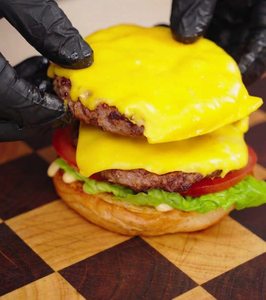 Hands in black gloves stacking two juicy Wagyu beef patties covered in melted cheese onto a burger assembly.