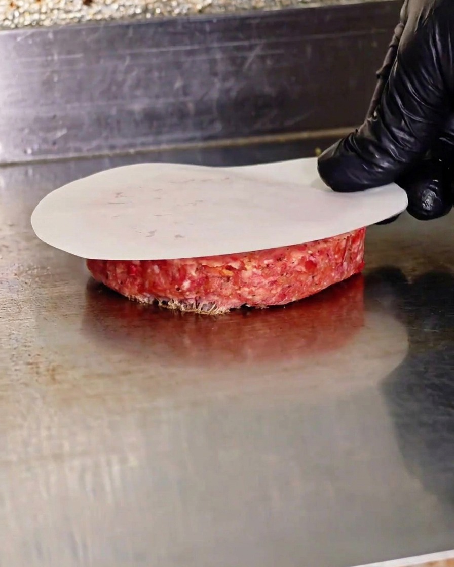 A gloved hand pressing down on a raw beef patty using a flat press over a piece of parchment paper on a hot griddle.