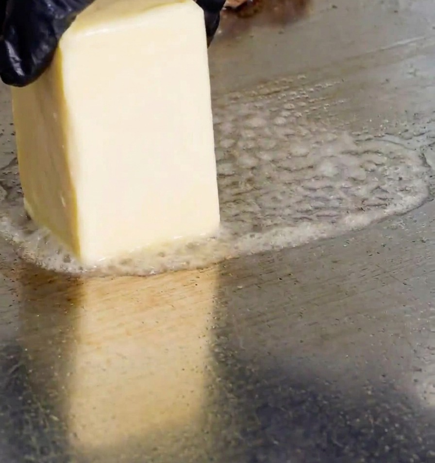 A hand wearing a black glove rubbing a rectangular block of butter onto a hot metal griddle, creating bubbles of melting fat.