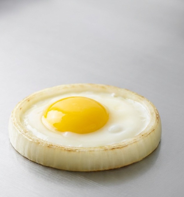 A perfectly circular fried egg with a bright yellow yolk cooking inside a thick onion ring on a flat stainless steel surface.
