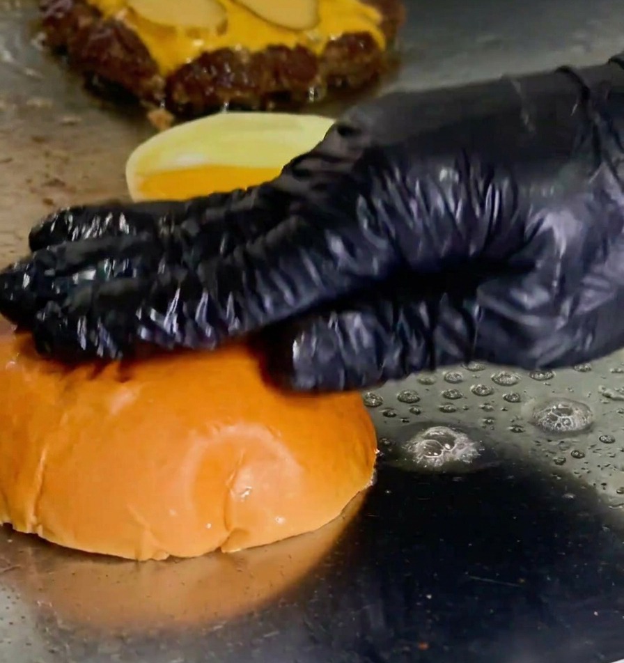 A gloved hand pressing a burger bun face-down into a pool of melted butter on a griddle next to a cooking patty.