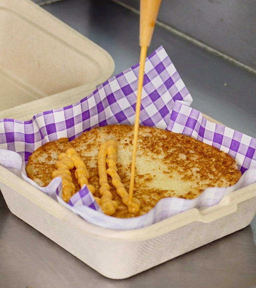 A stream of creamy orange sauce being squeezed from a bottle onto the center of a toasted burger bun in a purple checkered box liner.
