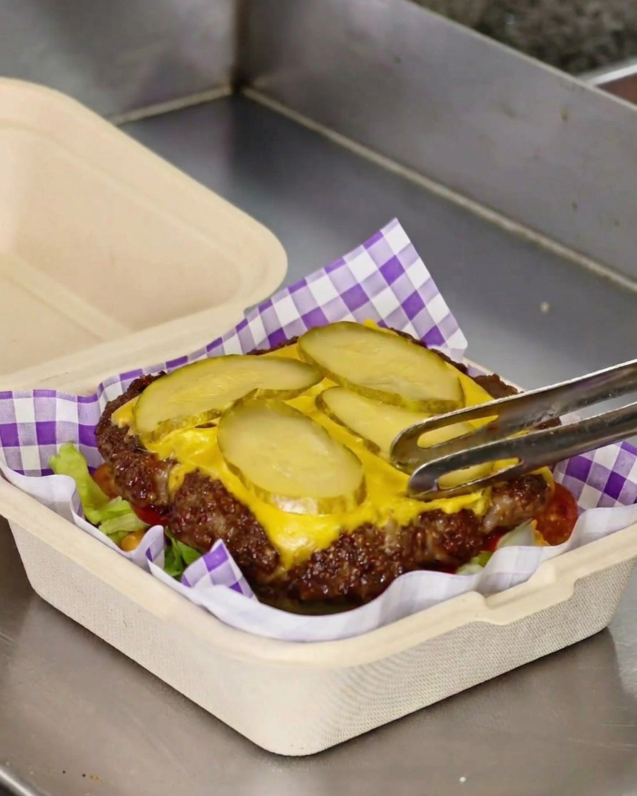 Tongs transferring a cooked smash burger patty topped with melted yellow cheese and pickle slices onto a bed of green lettuce.
